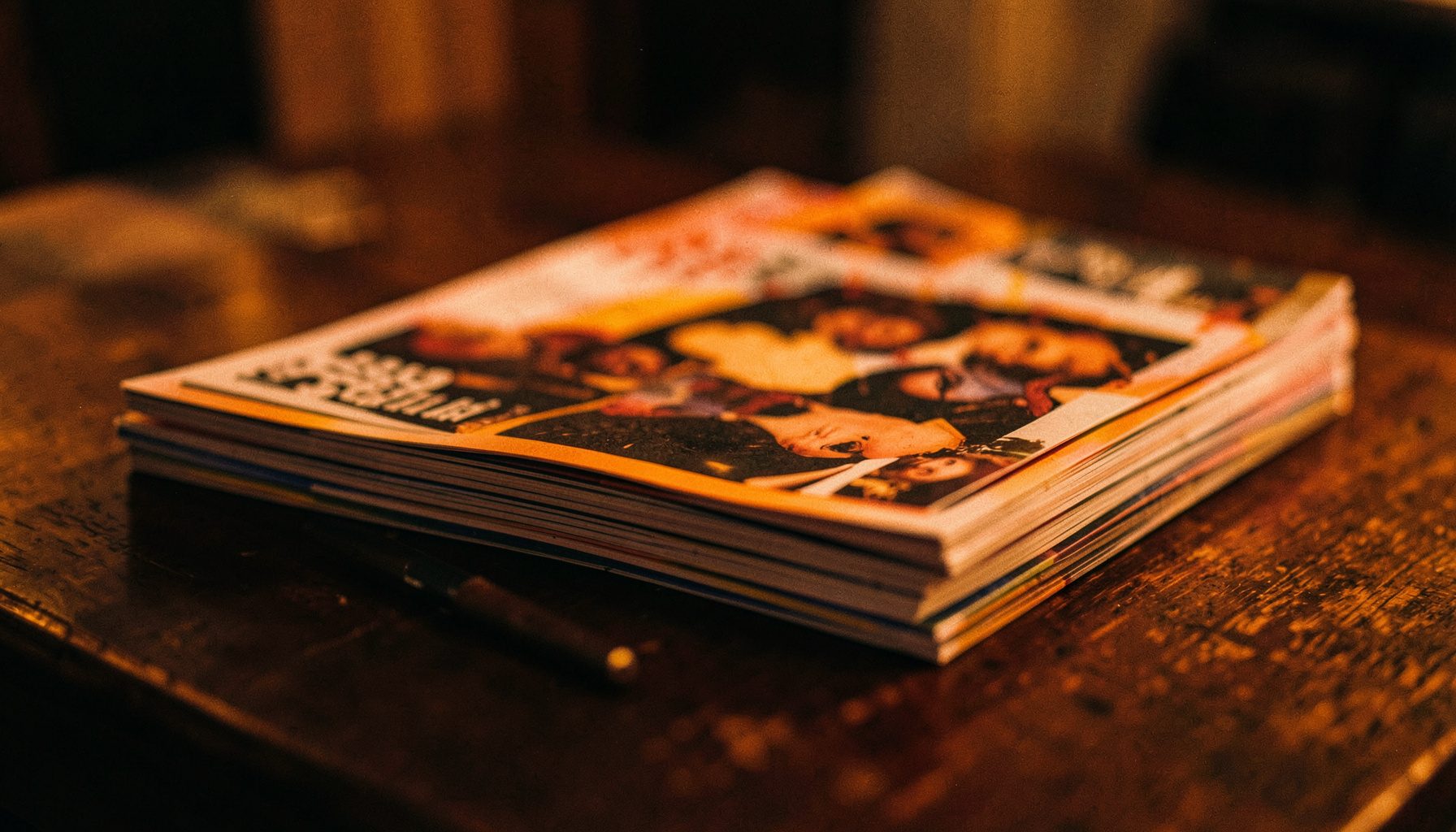 A stack of music magazines on a dark wooden table, warm amber and deep brown tones, shallow depth of field with soft shadows