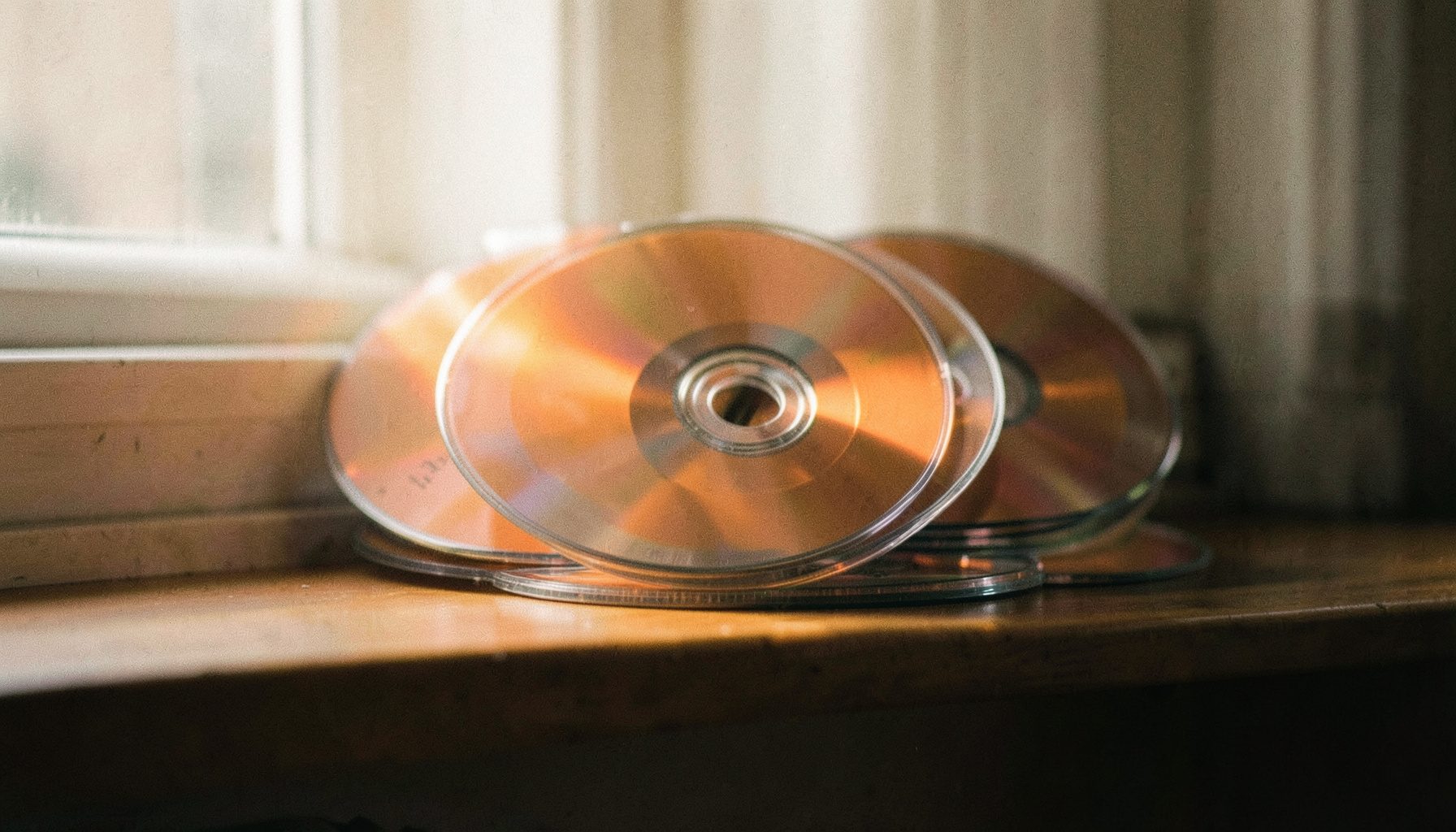 Compact discs stacked on a shelf with soft natural light from a window, muted tones