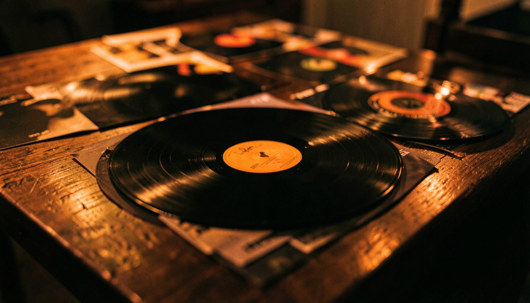 Vinyl records spread across a dark wooden table under warm tungsten light, shallow depth of field